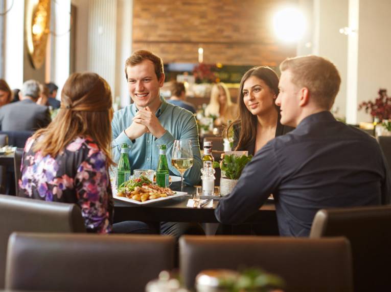 Vier junge Erwachsene sitzen in einem stilvollen Restaurant, unterhalten sich und genießen ein Essen bei Kerzenlicht.